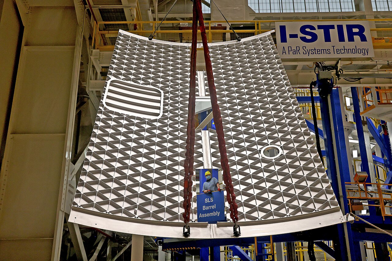 Curved orthogrid aluminium panel hoisted in the welding bay of a rocket factory.
