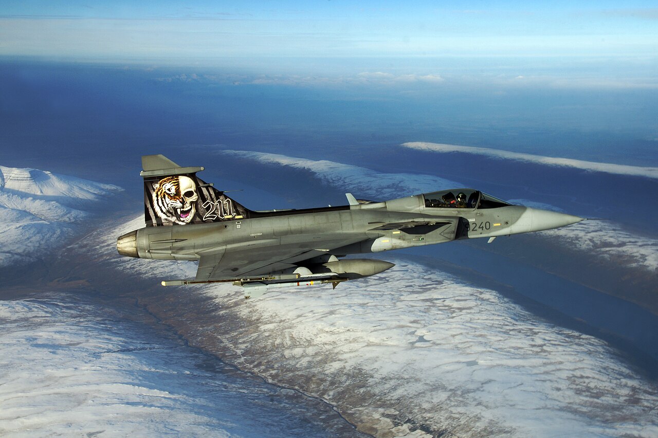 A Saab JAS 39 Gripen fighter in flight over a mountainous landscape.