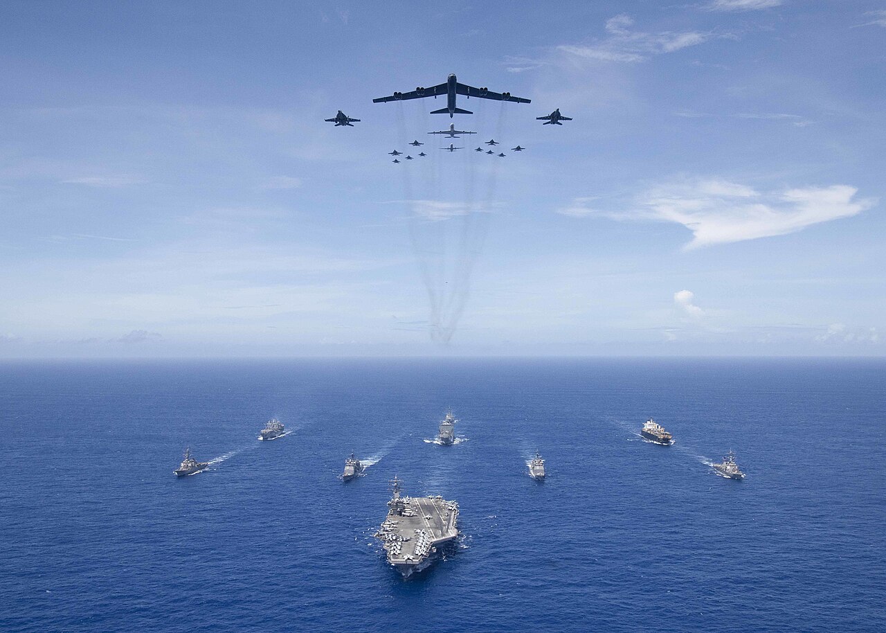 B-52 bombers and F/A-18 fighters fly in formation over an aircraft carrier strike group.